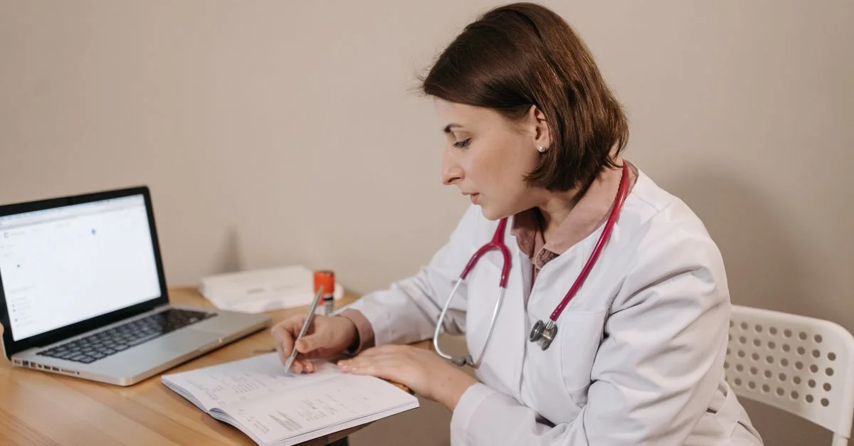 Une médecin portant une blouse d'hôpital écrivant une prescription dans un bureau clinique avec un ordinateur portable sur la table.