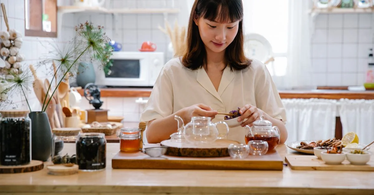 Femme préparant une infusion d'herbes avec des herbes et des épices dans une cuisine cosy baignée de lumière naturelle.